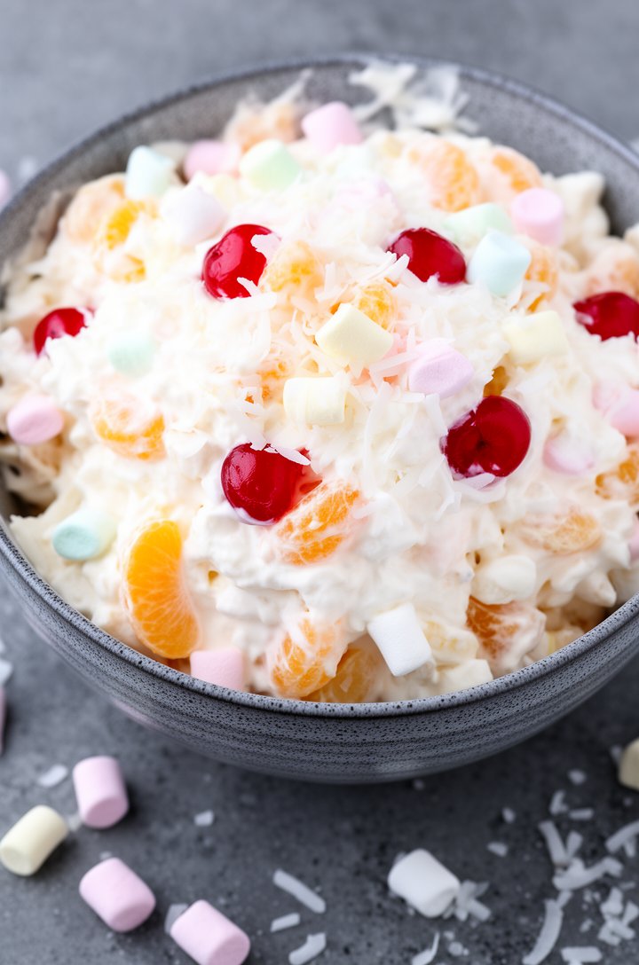 Extreme close-up overhead shot of finished ambrosia fruit salad mounded high in a gray speckled ceramic serving bowl, filling 90% of the frame. Fluffy white cream coating every piece — bright orange m