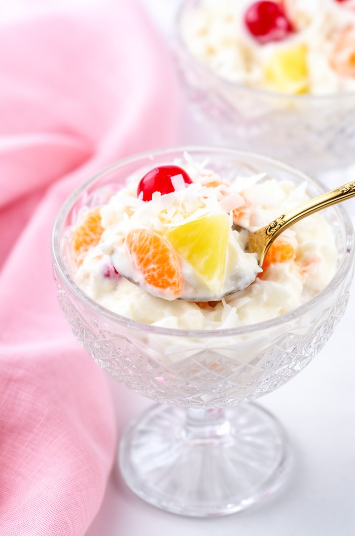 Close-up 30-degree angle shot of a single serving of ambrosia fruit salad in a crystal cut-glass dessert bowl, a gold vintage spoon lifting a generous scoop showing the creamy interior with visible ma