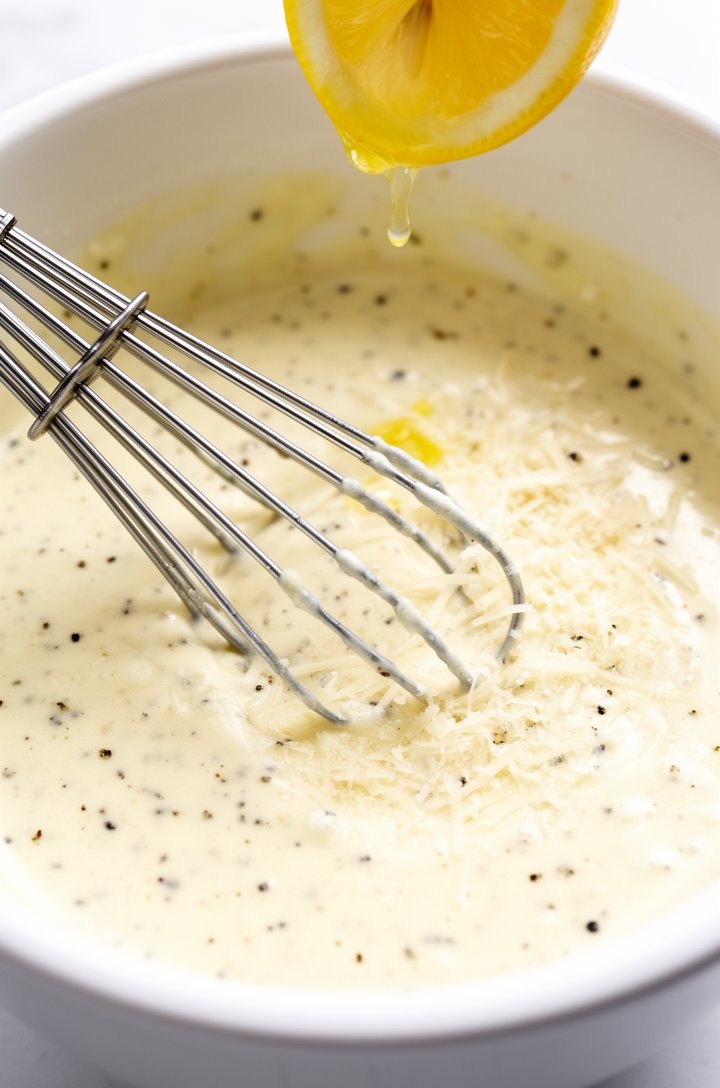 Extreme close-up of homemade Caesar dressing being whisked in a white ceramic bowl, thick creamy pale yellow texture with visible flecks of black pepper and grated Parmesan, a silver whisk resting in 