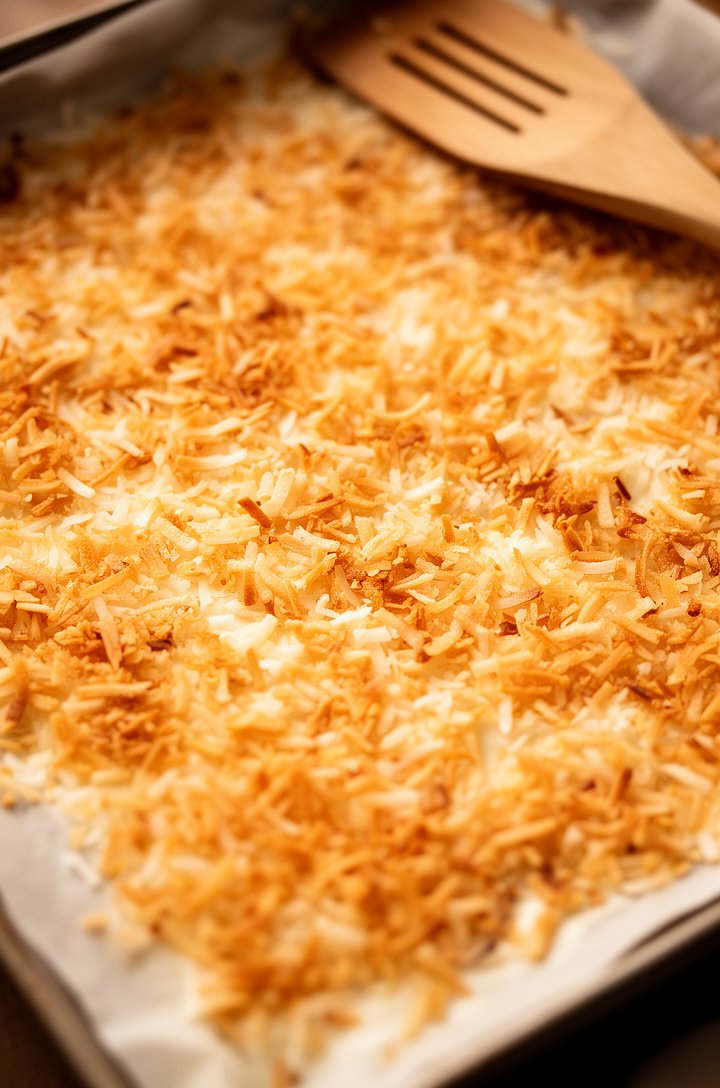 Overhead close-up of golden toasted coconut flakes spread on a parchment-lined baking sheet fresh from the oven, various shades from pale gold to deep amber, a wooden spatula resting on the edge, warm baking light