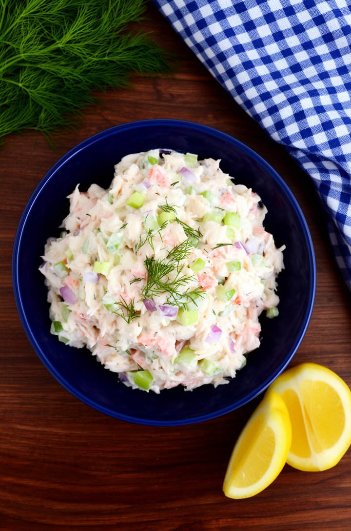 Overhead shot of finished creamy crab salad served in a deep navy blue ceramic bowl on a dark walnut wood table, the salad mounded generously showing chunks of white-pink crab coated in creamy dressin