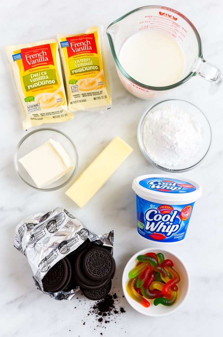 Overhead flat-lay of dirt cake ingredients arranged on a white marble surface — two packets of French vanilla instant pudding, a glass measuring cup of milk, a block of cream cheese, a stick of butter