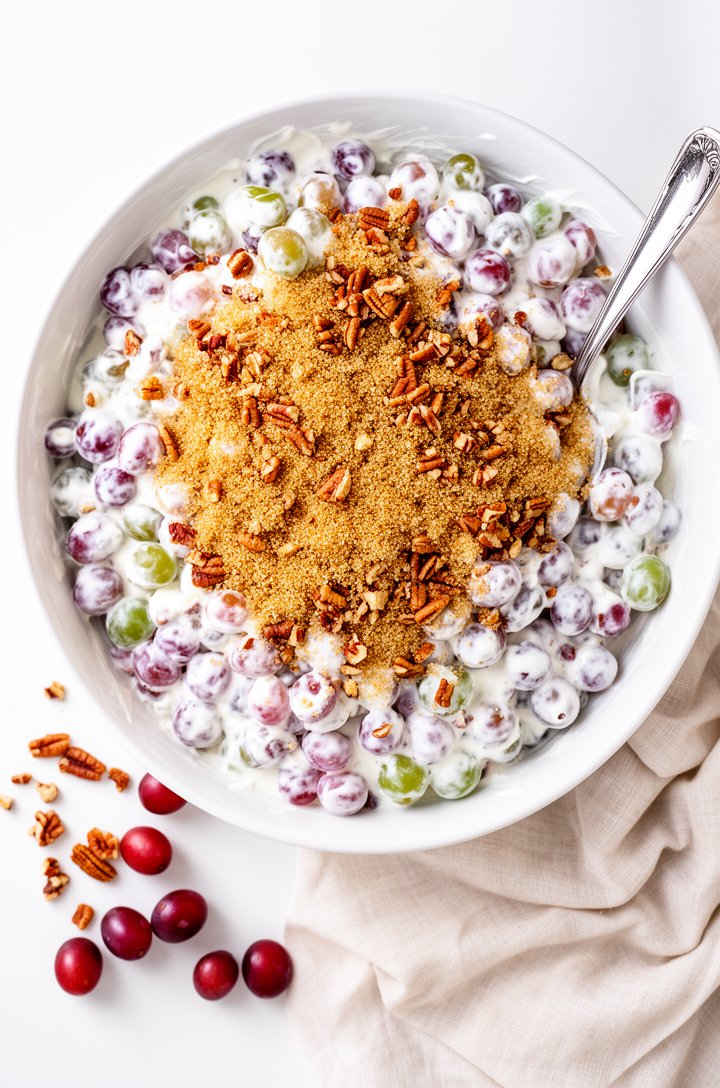 Overhead shot of finished grape salad in a large white ceramic serving bowl, mixed red and green grapes fully coated in thick white cream cheese dressing, generous crumble of golden-brown sugar and ch