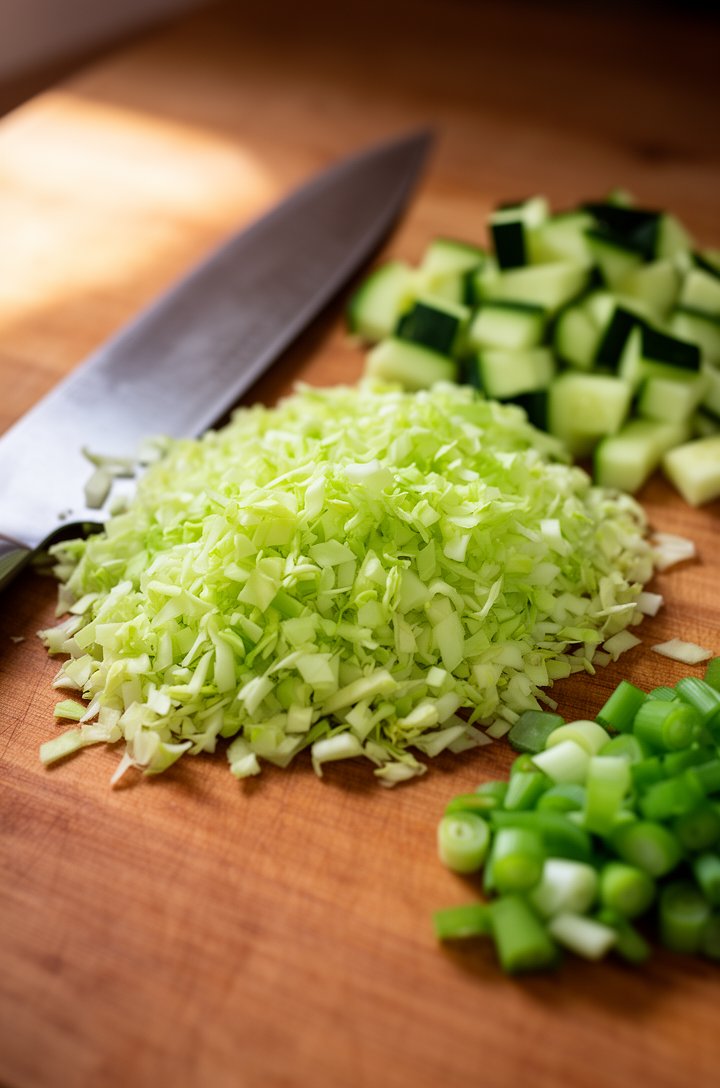 Close-up 45-degree angle shot of finely chopped confetti-sized green cabbage pieces on a wooden cutting board, a chef's knife resting beside the pile, with diced cucumber and sliced green onions in sm
