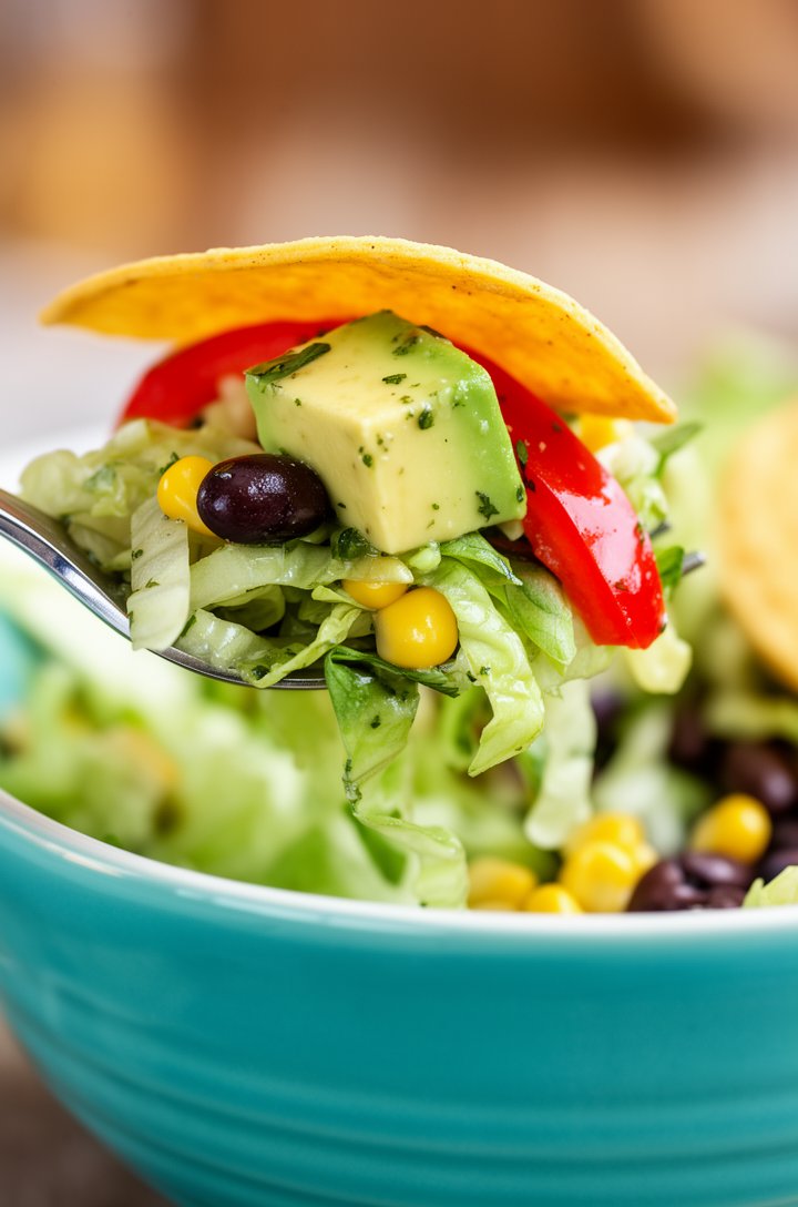 Extreme close-up side-angle shot of a fork lifting a generous bite from the Mexican salad, showing layers of chopped romaine, a black bean, a kernel of yellow corn, a cube of creamy avocado, and a sli