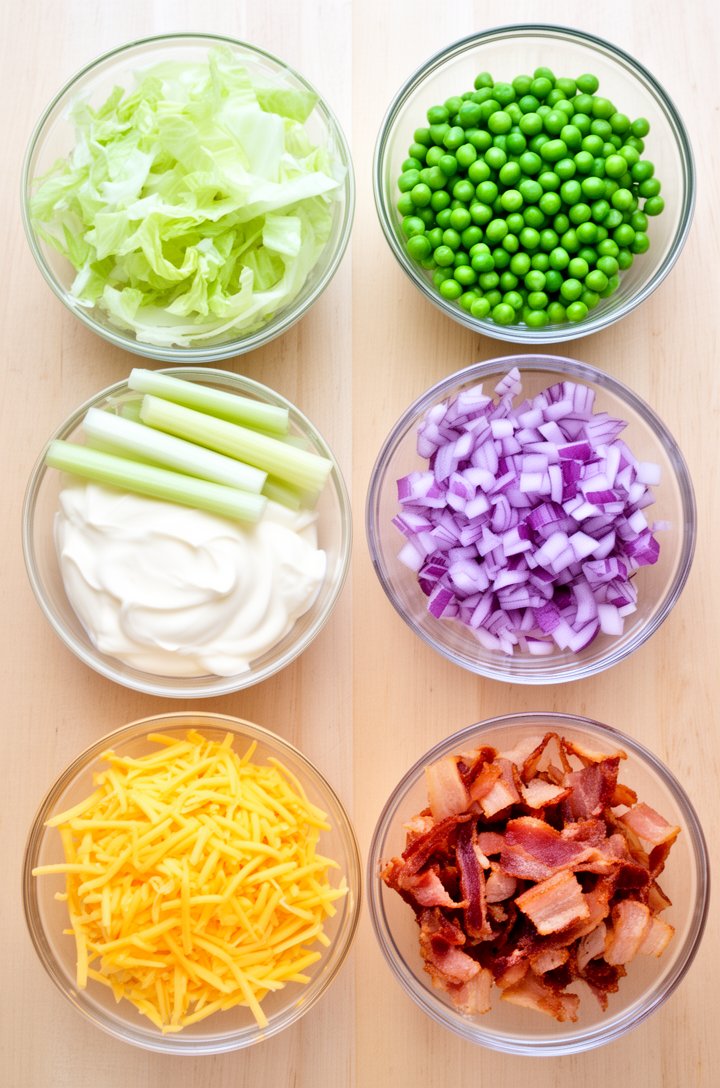 Overhead flat-lay of all seven layer salad ingredients arranged in small glass bowls on a light wood surface — a bowl of shredded iceberg lettuce, a bowl of bright green thawed peas, diced purple red 