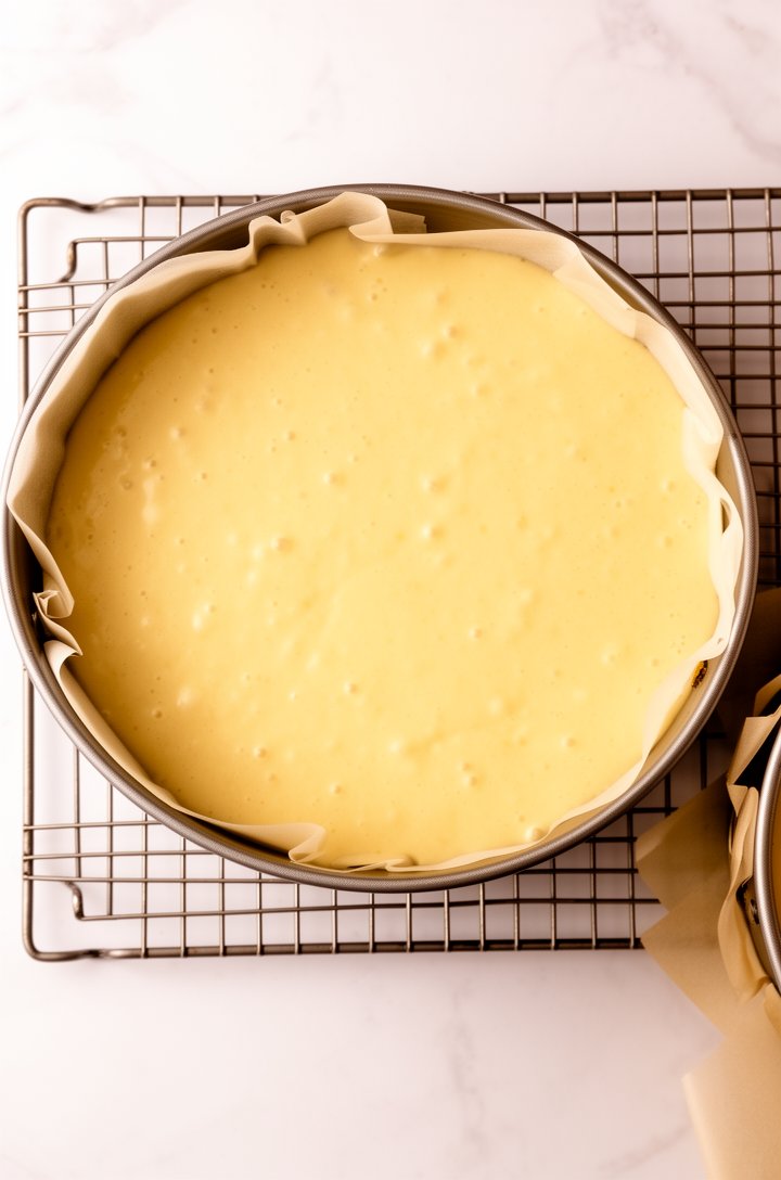 Overhead shot looking down into two round 8-inch cake pans filled with smooth pale yellow batter, sitting on a wire rack ready for the oven. The batter is pourable and glossy with tiny bubbles on the 